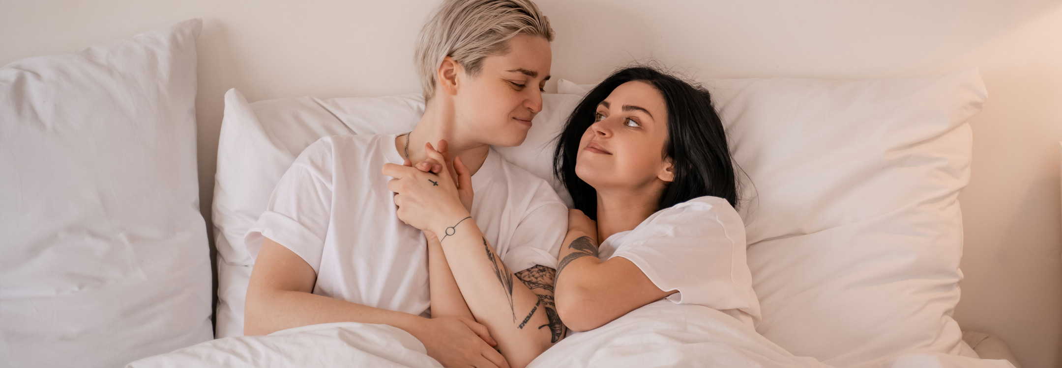 Two women lying down in bed with white clothes on looking at each other while holding hands